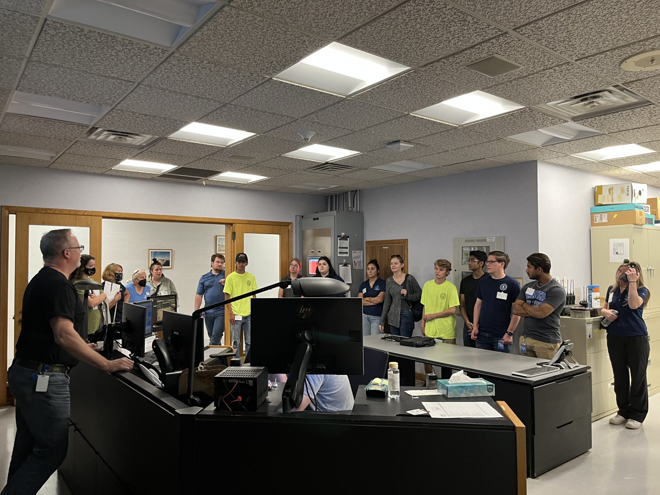 people gathered around a control center listening to a presentor